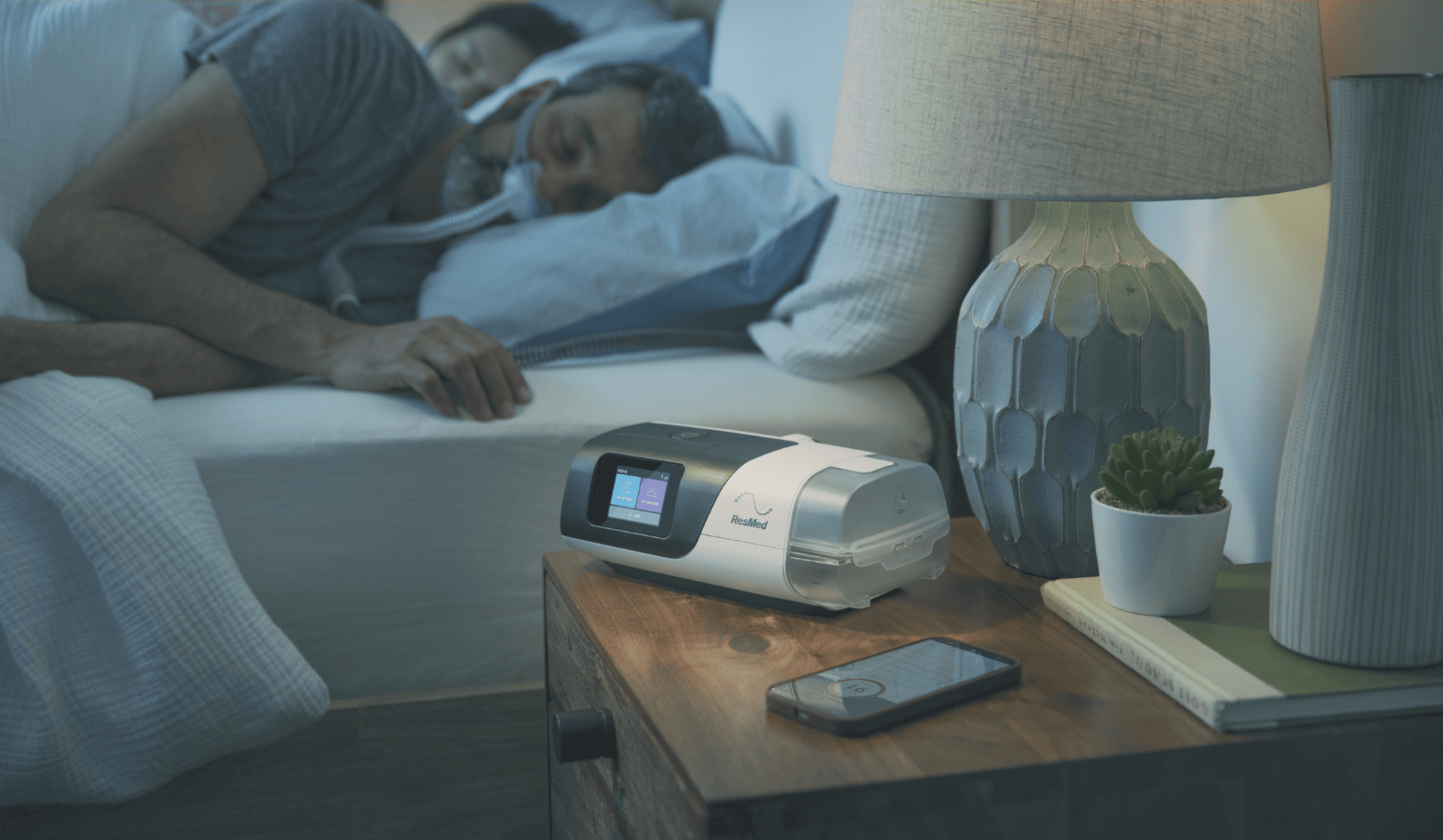 CPAP machine on nightstand beside sleeping person wearing a mask, improving sleep apnea treatment.